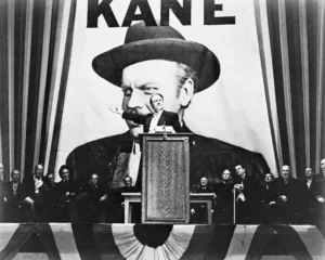Black and white image from the film "Citizen Kane," showing a scene where the character Charles Foster Kane, portrayed by Orson Welles, is giving a speech at a political rally