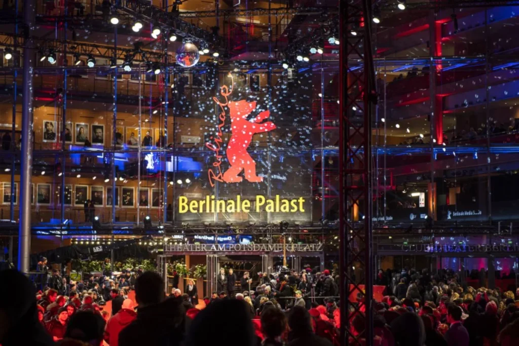 Crowd of people gathered at night outside the brightly lit Berlinale Palast, featuring a prominent red Berlinale bear logo