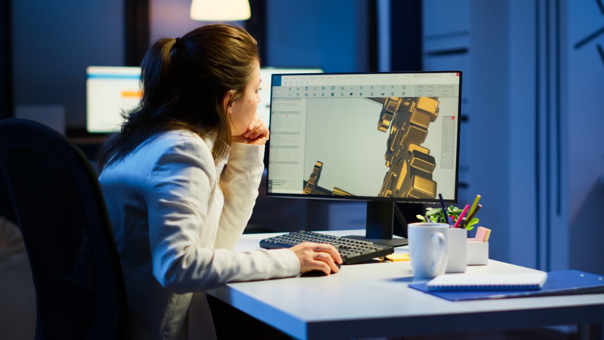 A woman in a white blazer is working late at a desk, focused on a computer displaying a 3D design, surrounded by office supplies.