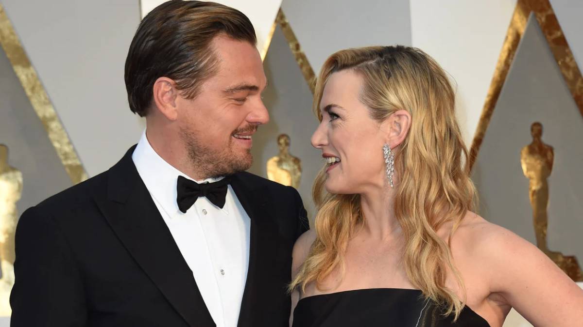 A glamorous couple poses together at an awards ceremony, with gold statuettes and a decorated backdrop in view.