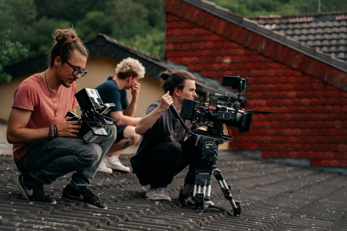 A group of filmmakers crouches on a rooftop, using various cameras and equipment to capture a scene, amid green trees and red-tiled roofs.