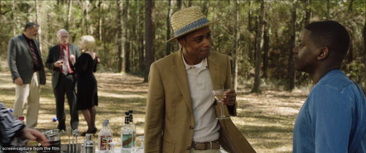 A gathering in a wooded area, featuring a man in a suit holding a martini and another in a denim shirt, surrounded by party guests.