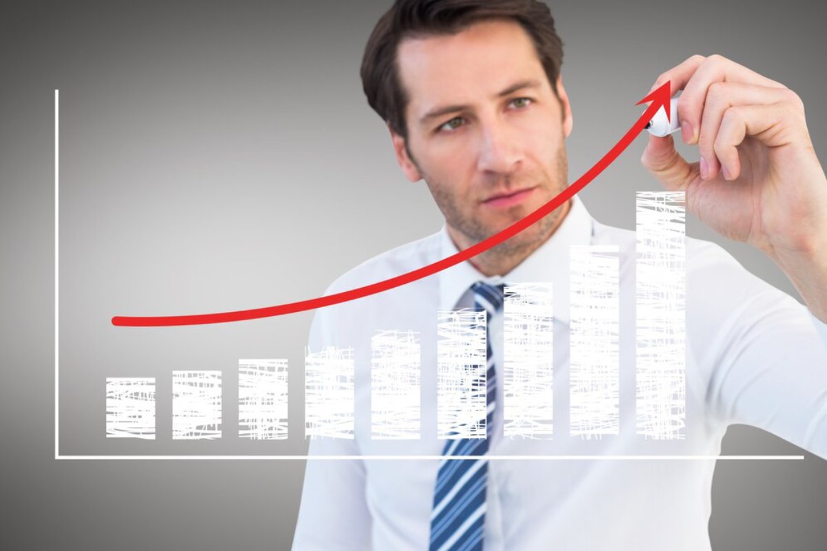 A businessman in a white shirt and tie draws a red upward trend line on a bar graph against a blurred background.