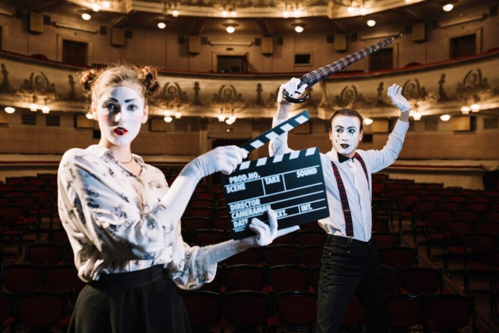 Two actors in a vintage theater, one holding a clapperboard and the other wielding a sword, both dressed in theatrical attire.