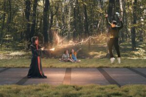 A wizard in a dark robe conjures sparks as another child leaps joyfully on a path, while two others sit and watch in a lush forest.
