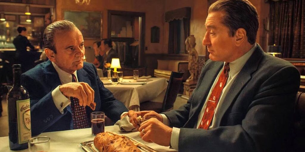 Two well-dressed men sit at a table in a dimly lit restaurant, sharing a conversation over drinks and food. A bottle of wine is visible.