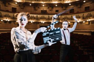 Two actors in a vintage theater, one holding a clapperboard and the other wielding a sword, both dressed in theatrical attire.